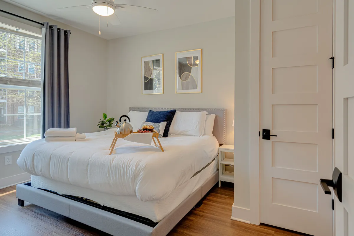 A serene bedroom scene with a plush bed, a breakfast tray featuring tea and snacks, soft artwork on the wall, and daylight streaming in. The overall feeling is one of calm and comfort, inviting relaxation and leisurely mornings in bed.