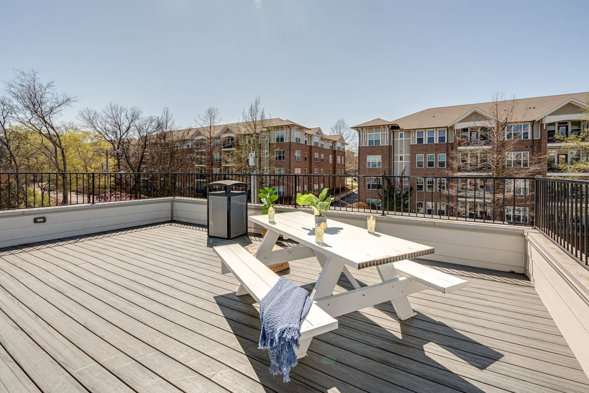 An inviting rooftop deck featuring an outdoor dining table surrounded by comfortable chairs and lush planters filled with greenery. The space is open to the sky, framed by views of nearby residential buildings. The ambiance is bright and cheerful, suggesting both daytime and evening usability.