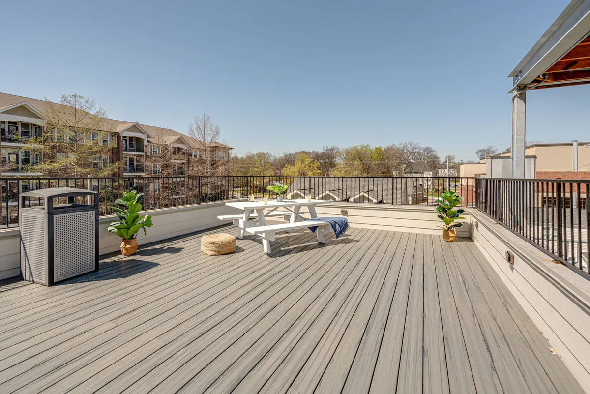A casual outdoor patio with a picnic table set among green plants. The surrounding area is relaxed and welcoming, offering space for dining or gathering with friends. A trash can and fence indicate a functional, lived-in urban setting.