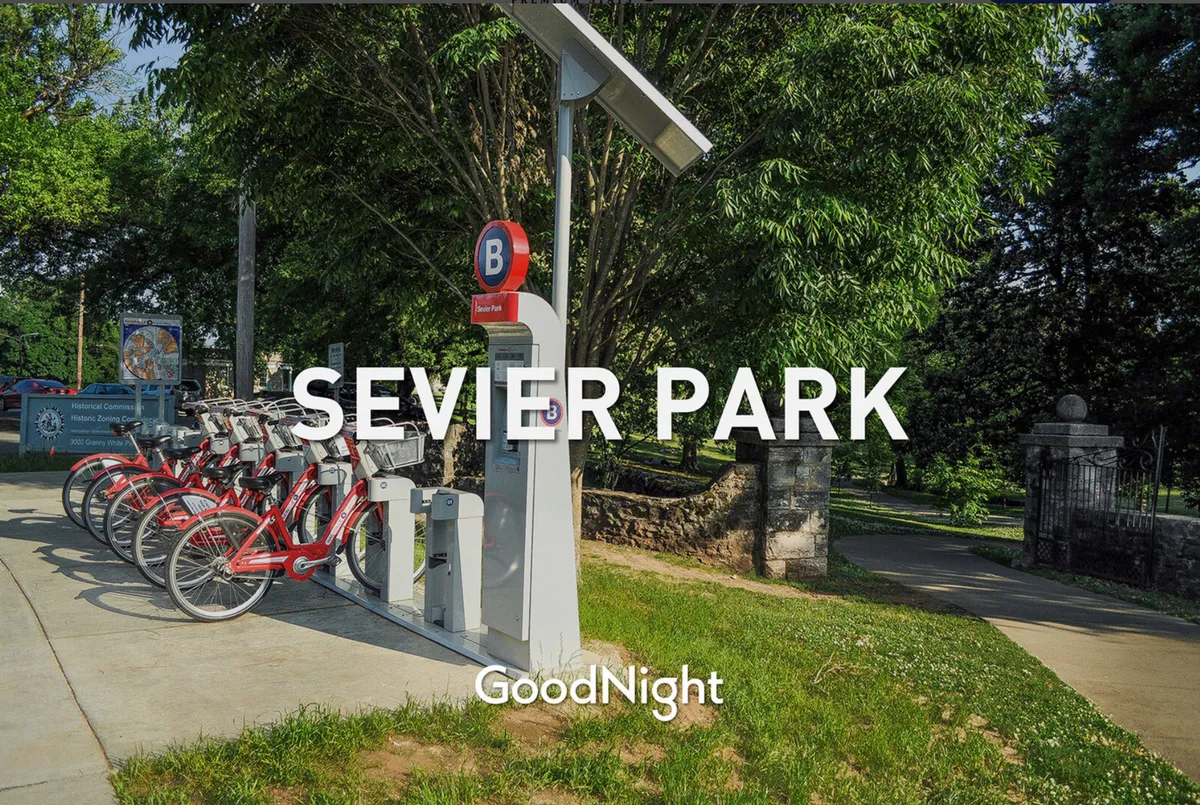 A sunny outdoor view of Sevier Park with a bicycle rental station, the Sevier Park sign, and ample green surroundings. People are seen biking and walking, highlighting the area's active and family-oriented vibe.