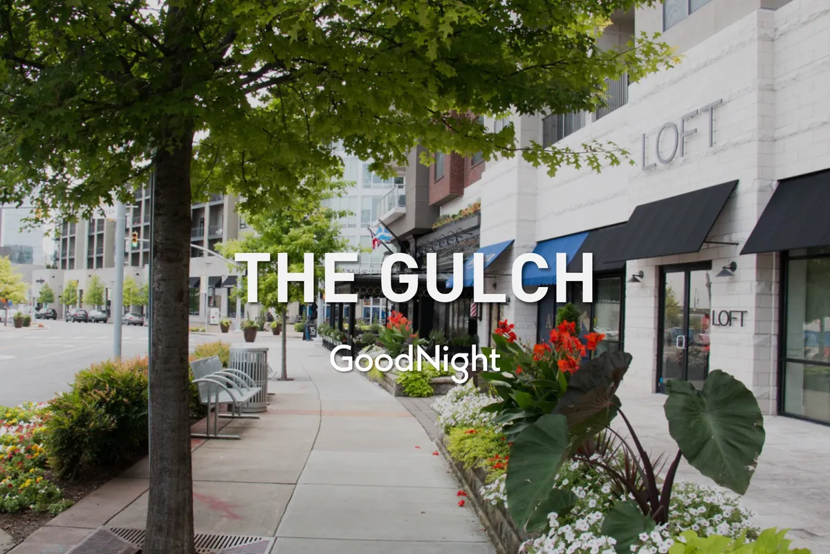 A lively Nashville street scene featuring tree-lined sidewalks, colorful flower beds, storefronts, and benches. Locals and visitors enjoy walking, shopping, and relaxing outdoors, capturing the neighborhood's friendly, walkable atmosphere.