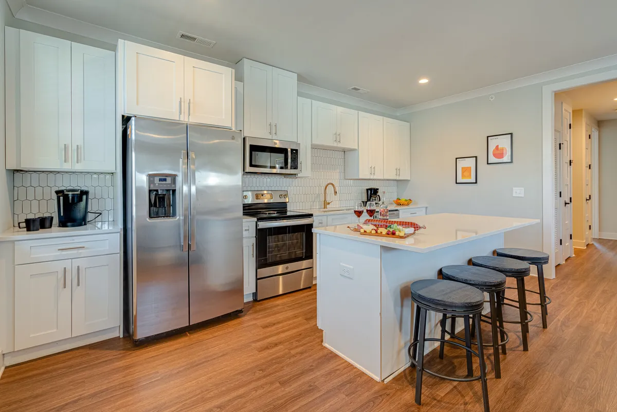 The image showcases a bright, modern kitchen with gleaming stainless steel appliances, a spacious island countertop with stylish bar stools, a decorative fruit bowl, and tasteful framed art on the walls. The space is inviting and perfect for both cooking and gathering, exuding a welcoming, contemporary vibe.