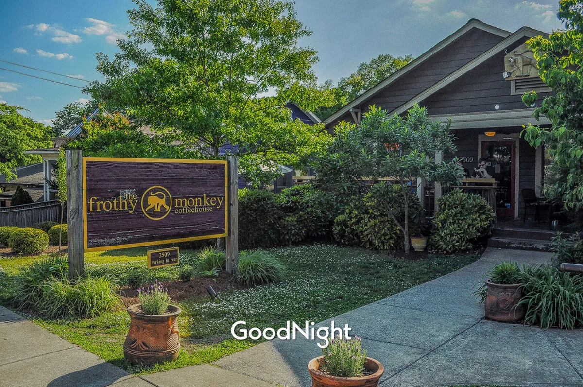 The entrance of Frothy Monkey coffeehouse, surrounded by lush greenery and inviting landscaping. The scene feels relaxed and welcoming, with a visible sign and cozy outdoor area, hinting at the vibrant local dining and café culture.