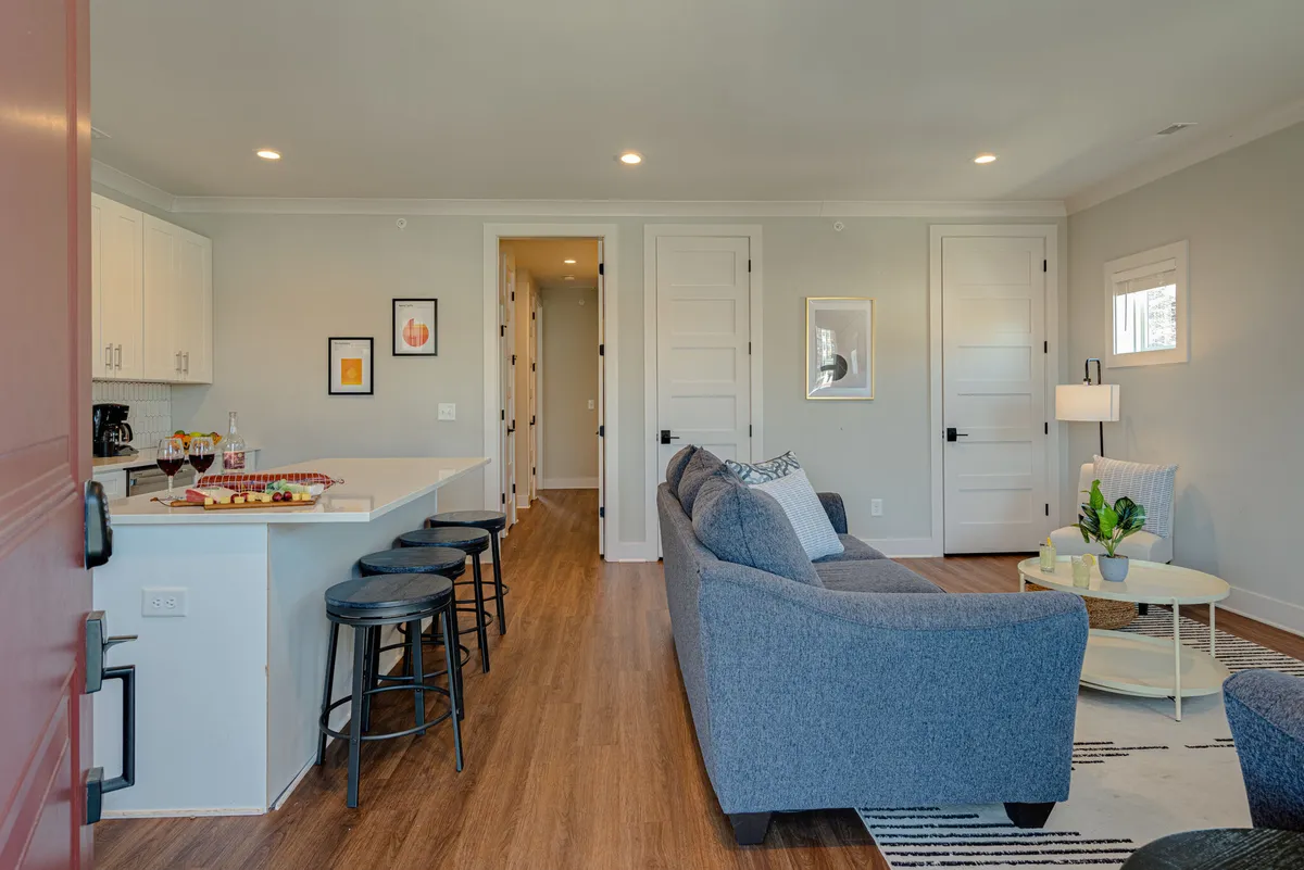 The image presents a seamless integration of a modern sofa and a kitchen island with wine glasses, accented by colorful artwork on the wall in the background. The open layout subtly connects the living and kitchen areas, highlighting the space's sociable and multifunctional design.