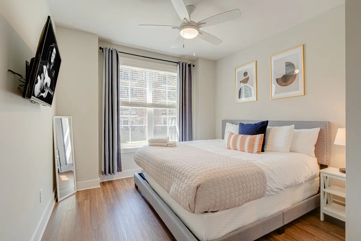 A tranquil bedroom scene featuring a king-size bed adorned with plush bedding, a window allowing natural light to flow in, tasteful wall artwork, and a mounted television. The setting exudes calm, with soft lighting and a cozy atmosphere perfect for restful sleep or relaxed mornings catching up on shows.